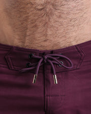 Close-up view of men’s summer beach shorts by BANG! clothing brand, showing dark purple cord with custom branded golden cord ends, and matching custom eyelet trims in gold.