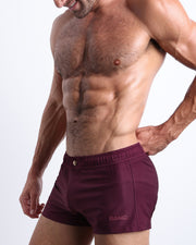 Side view of a masculine model wearing the men’s VERY BERRY shorter leg length swim shorts in red wine color with official logo of BANG! Brand in a matte light burgundy color.