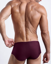 Back view of a male model wearing men’s swim sungas in a neon highlighter dark wine red color by the Bang! Clothes brand of men's beachwear.