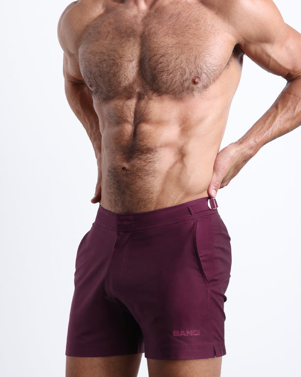 Side view of a masculine model wearing men’s swimsuit shorts in VERY BERRY a dark purple/red color featuring a side pocket with official logo of BANG! Brand in a matching purple color.