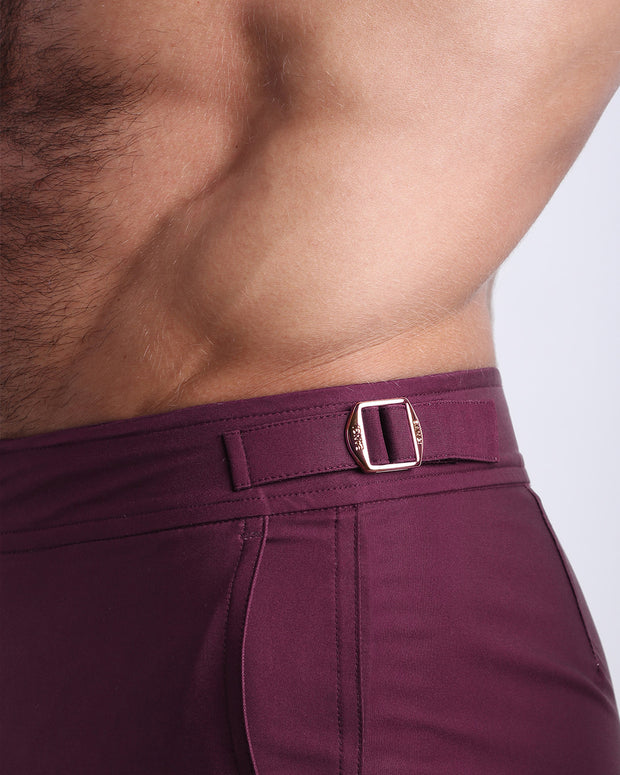 Side view of a masculine model wearing men’s swimsuit shorts in VERY BERRY a dark purple/red color featuring adjustable metal side fasteners for tunable fit around waistline, with custom BANG! brand engraving.
