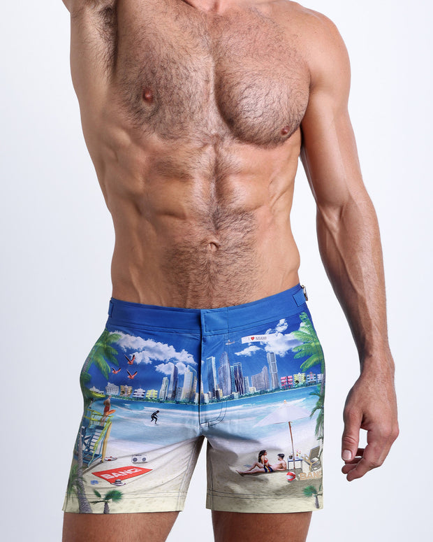 Front view of model wearing the WISH YOU WERE HERE men’s beach tailored shorts in a sky blue color featuring the Miami Beach skyline by the Bang! Clothes brand of men&