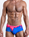 Frontal view of a sexy male model wearing men’s Brazilian Sunga swimsuit in YOU MELT ME in a bright blue color featuring pink melting ice cream print by the Bang! Menswear brand from Miami.